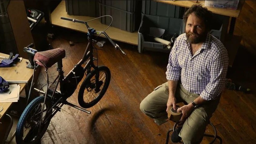 A bearded man sits next to a bicycle and a workbench, smiling.