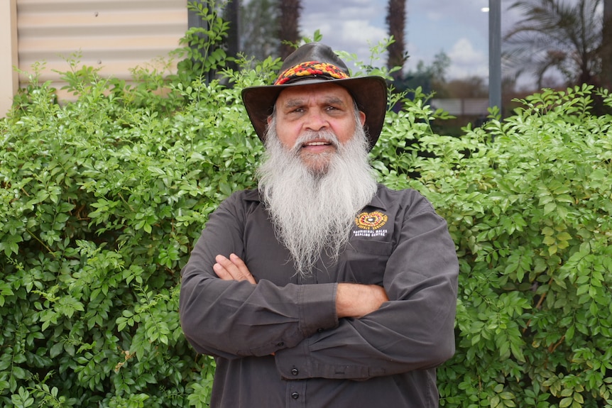 A man wearing a hat with a long white beard smiles in front of a green hedge.