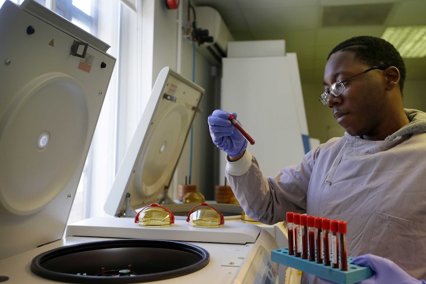 A man wearing white in a lab looks at a test tube in front of what looks a bit like an open washing machine.