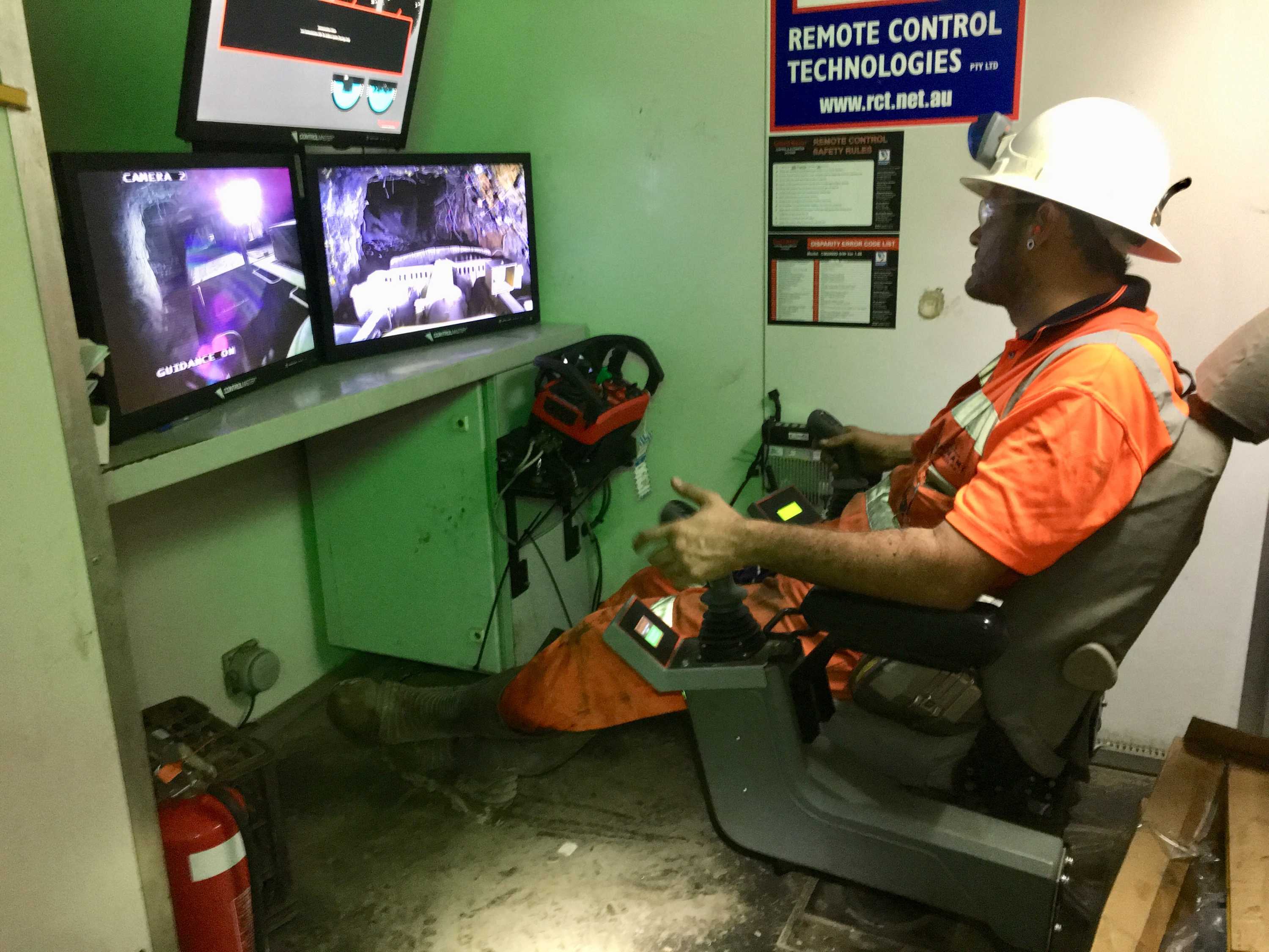A worker sits in high vis with remote controls in front of screens to control mining trucks remotely