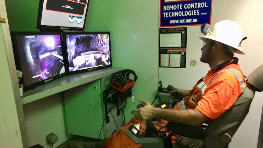 A worker sits in high vis with remote controls in front of screens to control mining trucks remotely