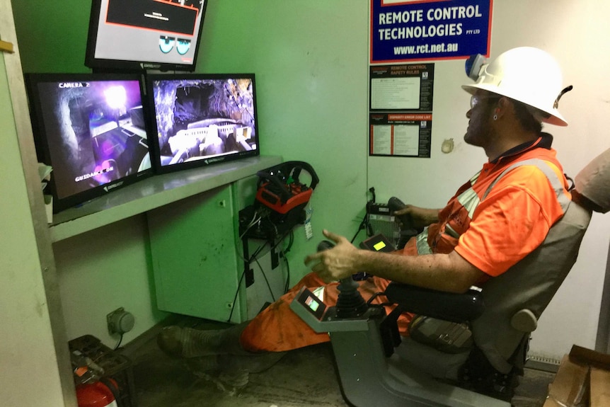 Un trabajador se sienta en alta visibilidad con controles remotos frente a pantallas para controlar camiones mineros de forma remota