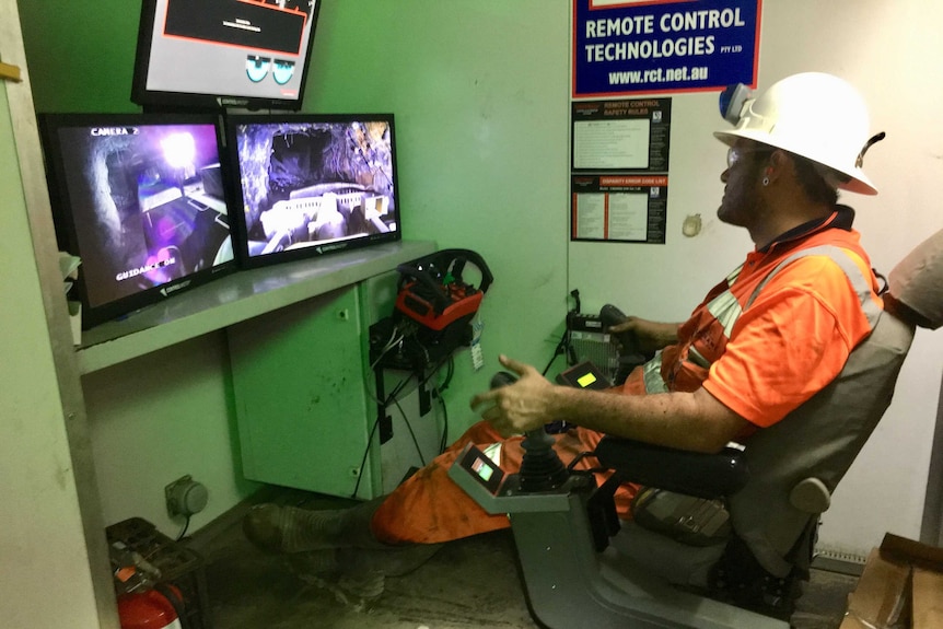 A worker sits in high vis with remote controls in front of screens to control mining trucks remotely