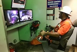 A worker sits in high vis with remote controls in front of screens to control mining trucks remotely