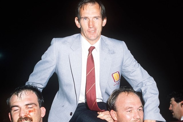 A middle-aged man in light jacket and suit & tie is carried from field by two rugby players in red.