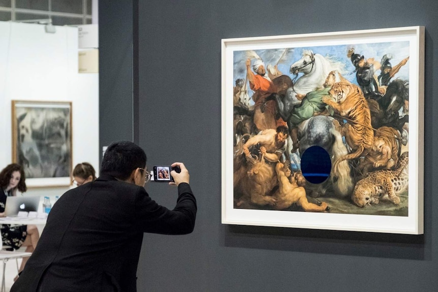A man takes a phone of a painting of men and animals wrestling.