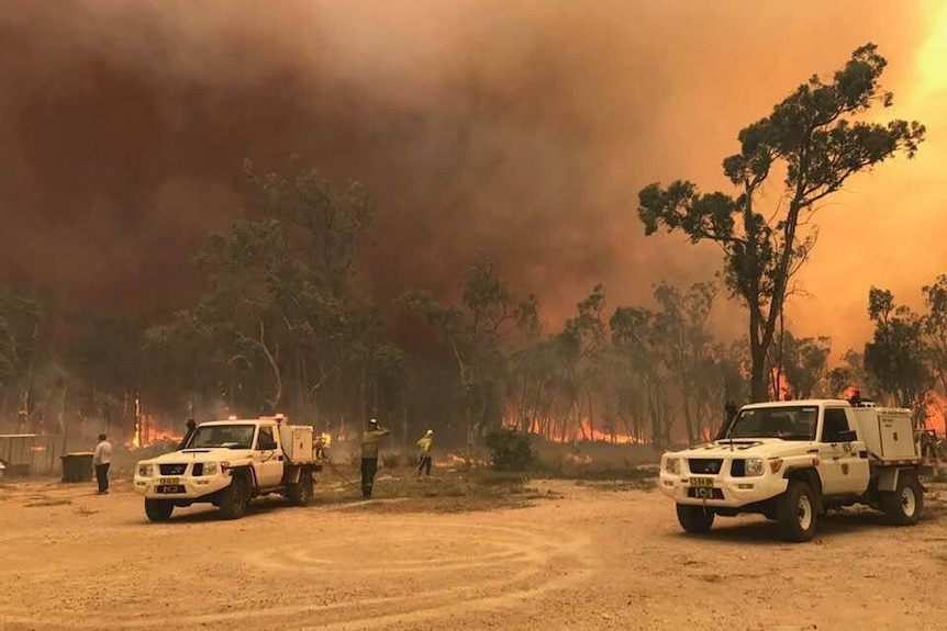 Three firefighters are seen battling fire with hoses as blaze rages in the background, with orange skies overhead.