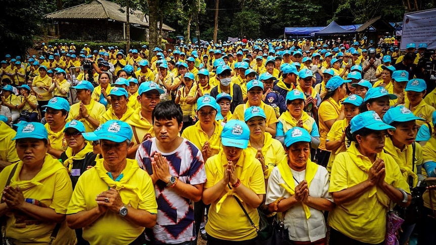 Scores of people dressed in yellow shirts and blue caps stand together, many with their hands pressed together