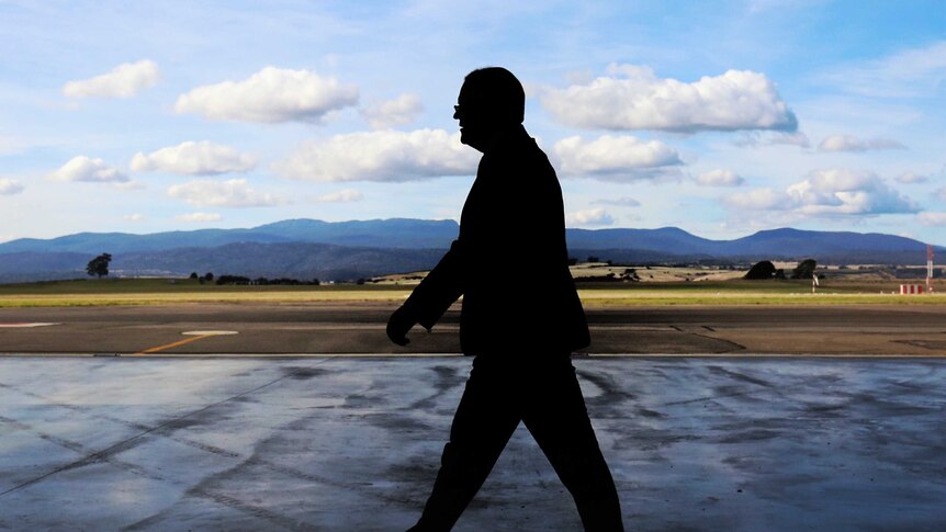 Mr Morrison, in shadow, walks through an airport hanger with mountains in the backgrounds