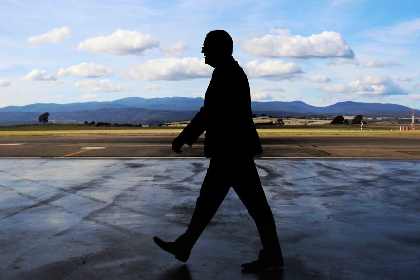 Mr Morrison, in shadow, walks through an airport hanger with mountains in the backgrounds