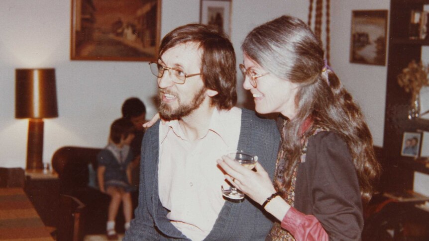 An old photo of a couple smiling in a lounge room, holding drinks.