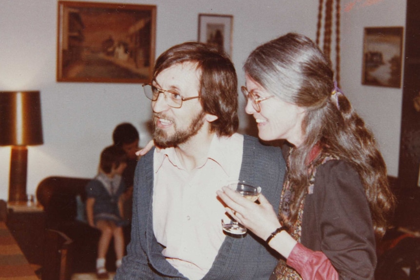 An old photo of a couple smiling in a lounge room, holding drinks.