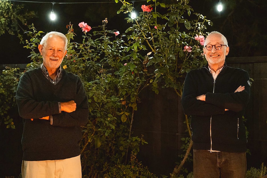 Two grey-haired men stand with arms crossed in front of greenery