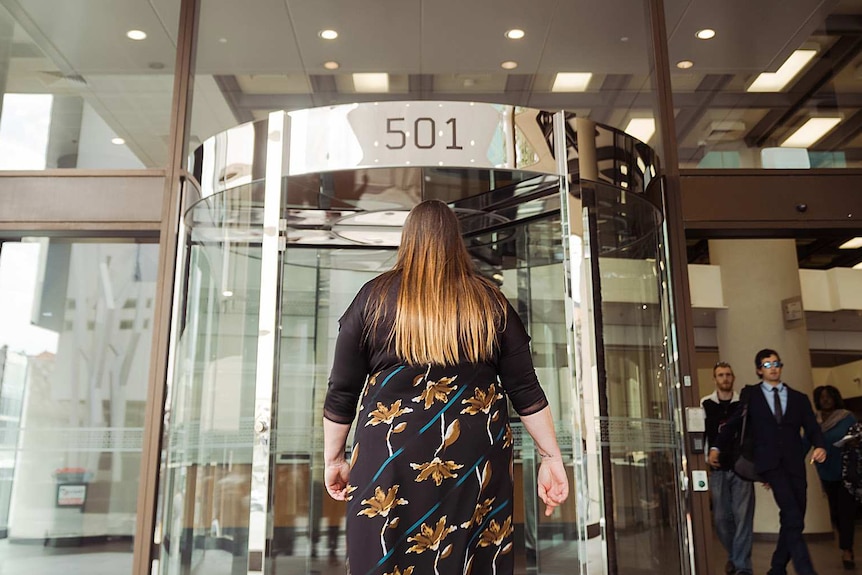 A woman in a black dress with flowers approaches a revolving glass door at the front of a court building.