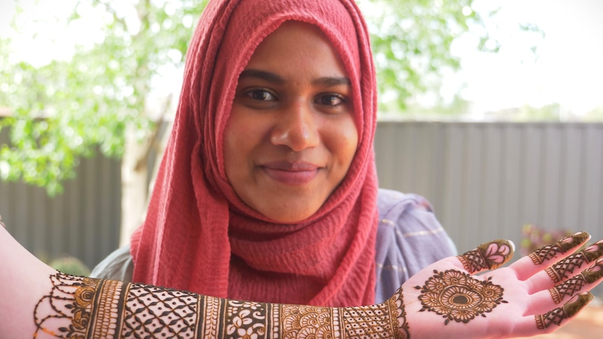 A woman in a hijab smiling to camera, a forearm covered in henna designs below her face