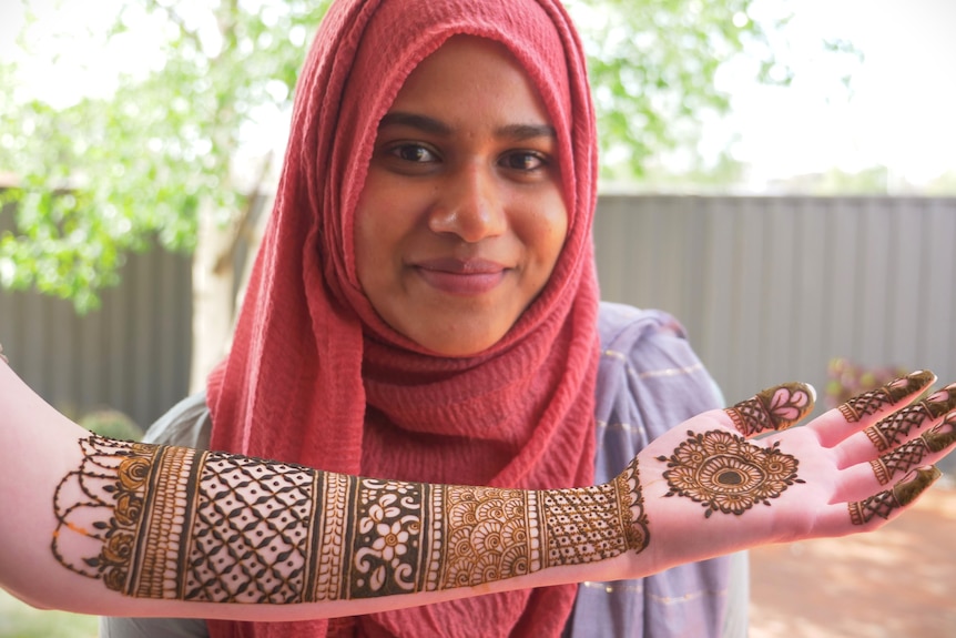 A woman in a hijab smiling to camera, a forearm covered in henna designs below her face