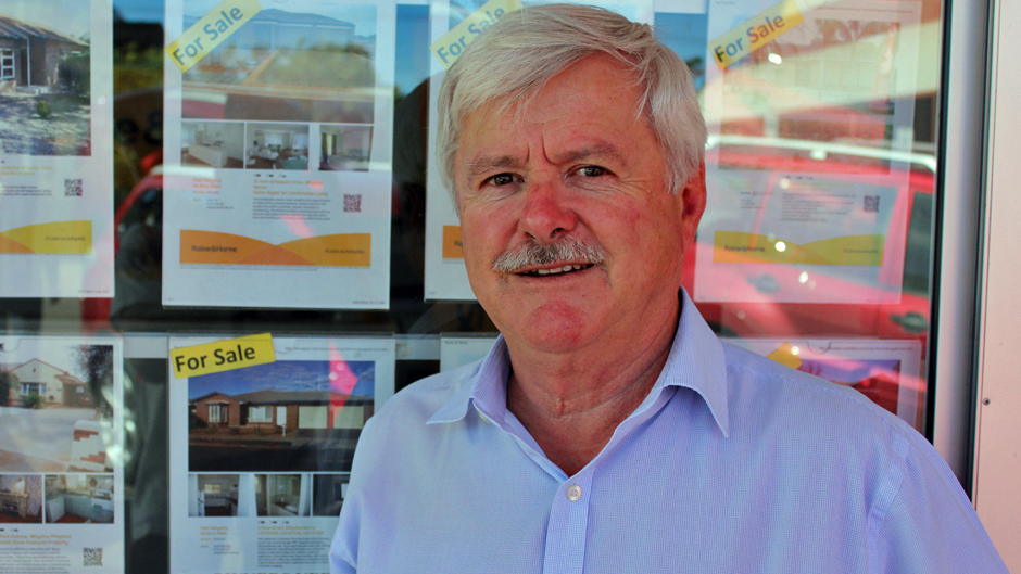 A man with grey hair and moustache and a blue shirt standing in front of real estate for sale signs.