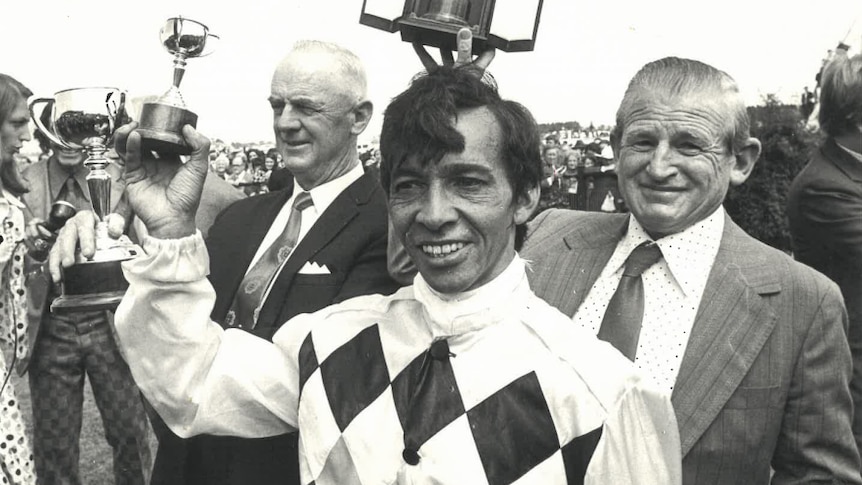 A black and white photo of a jockey holding a trophy