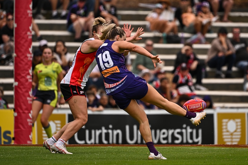 A Fremantle AFLW player kicks at goal as a St Kilda defender tries to smother.