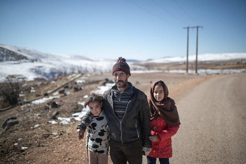 A man and his daughters pose for a photo with a wide, snowy landscape behind them.