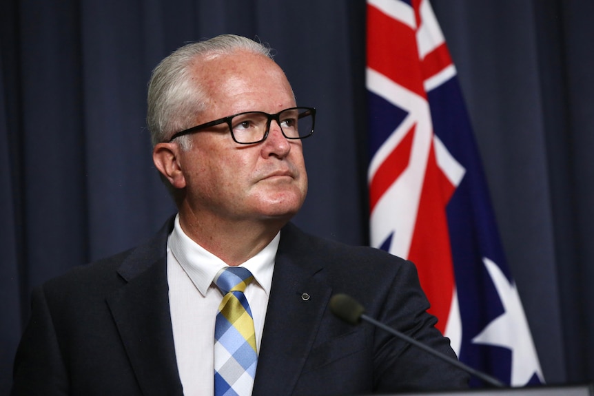 An older, bespectacled man with short white hair in front of an Australian flag.