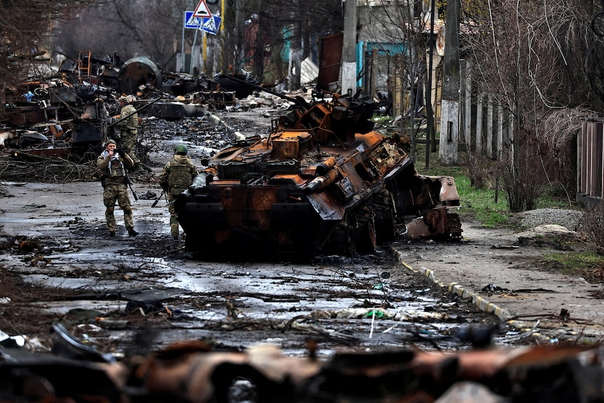 Two soldiers stand next to a blackened tank in a street strewen with blackened damage