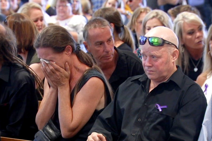 A woman holds a tissue to her eyes while other people look upset at a funeral.