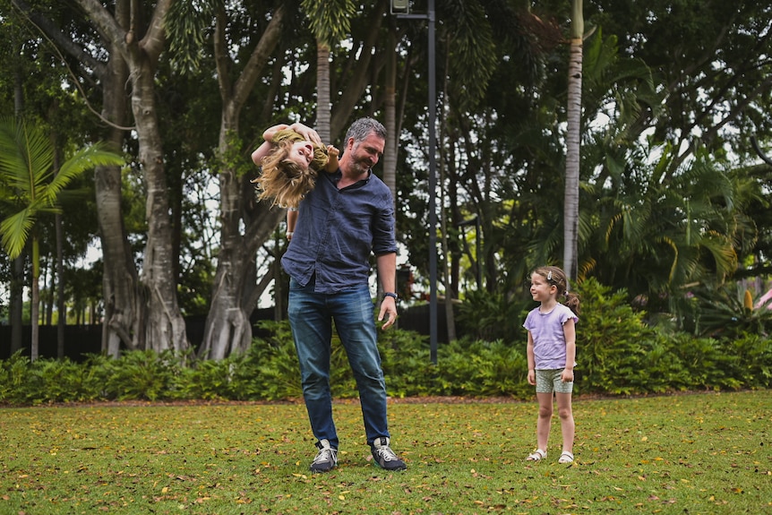 One little girl over her dad's shoulder, with another girl looking and smiling.