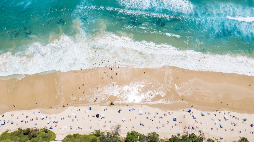 An aerial view of a beach with turquoise water, waves and sun bathers and swimmers
