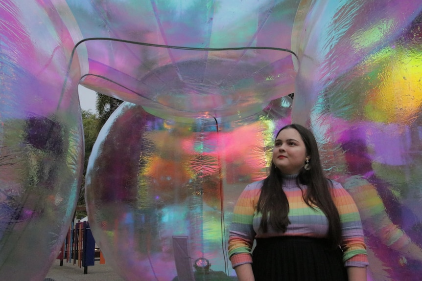 a woman with long brown hair wearing a colourful top inside a rainbow sculpture.