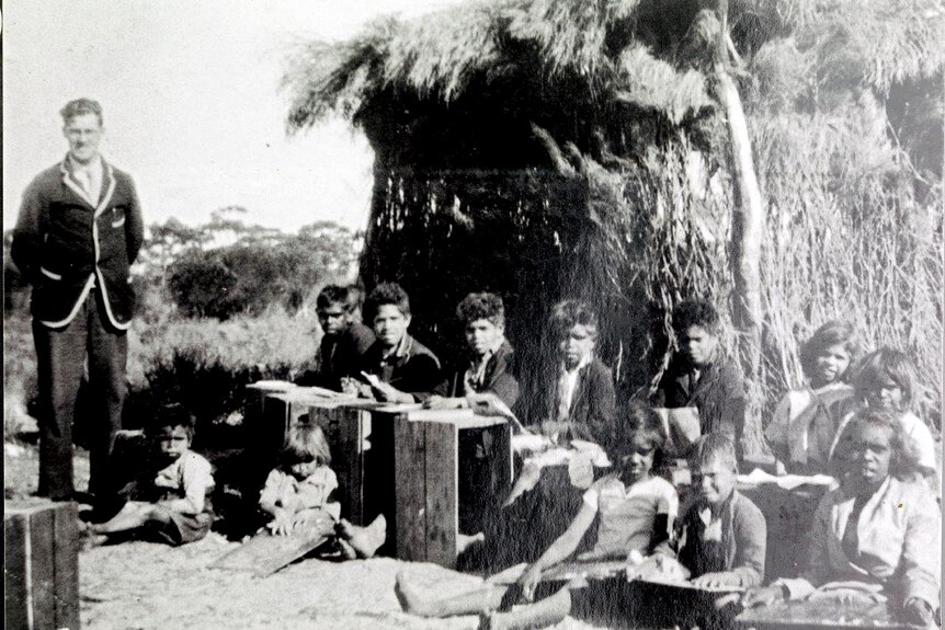 Children with work books sit on the ground or behind wooden crates as a man in a blazer stands on the side.