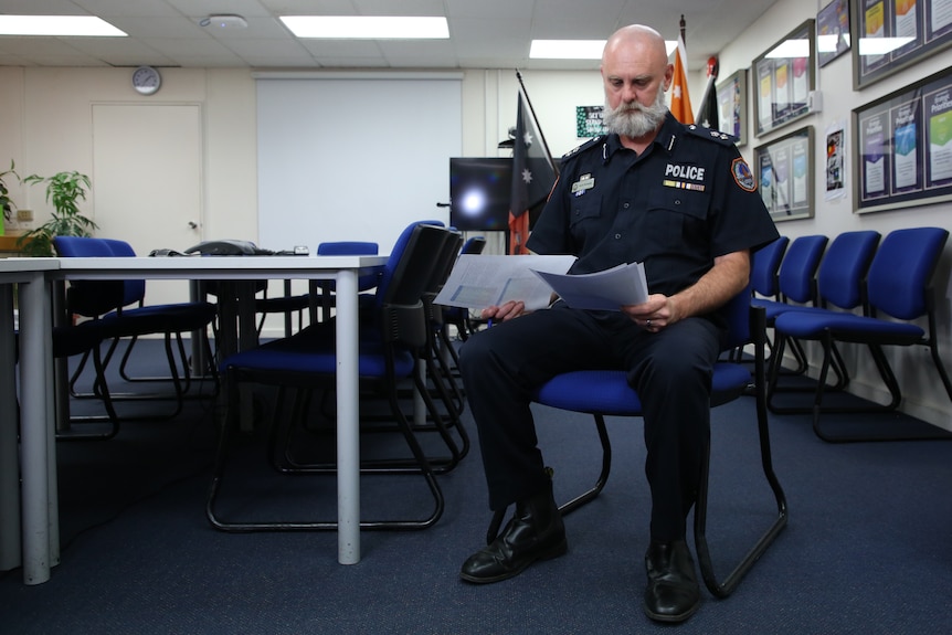A bald police officer sits in a blue chair, looking at paperwork.