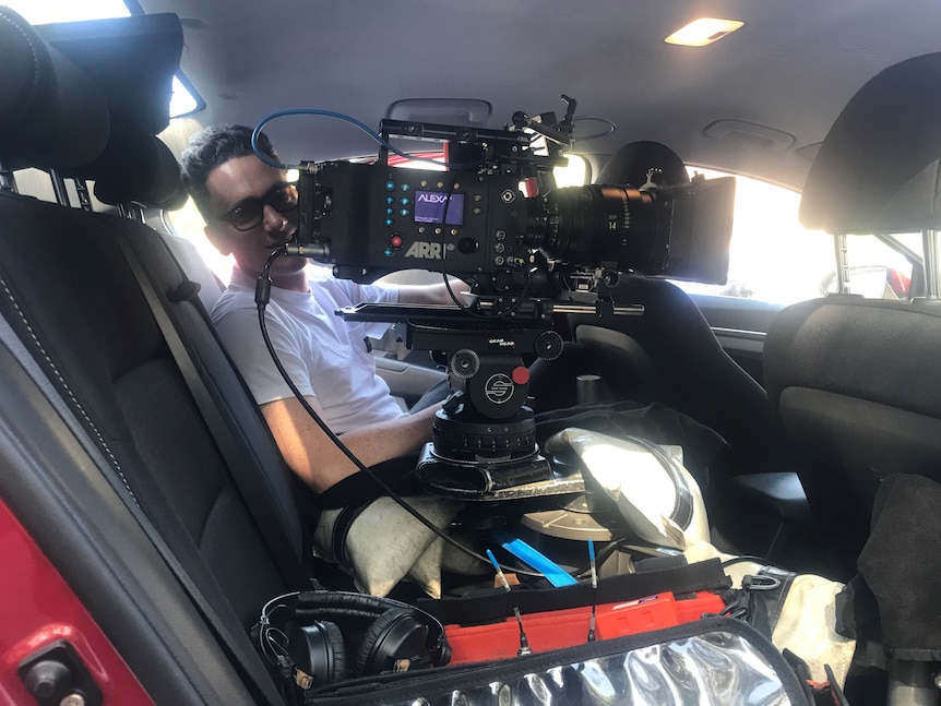 A man sitting next to a cinema camera in the backseat of a car.