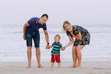 A woman and a man hold the hands of a small boy as they stand on sand in front of a calm beach.