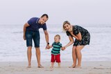 A woman and a man hold the hands of a small boy as they stand on sand in front of a calm beach.