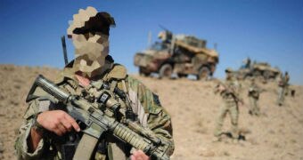 A soldier, whose face is pixelated, stands in camouflage gear with a firearm. In the distance behind him are trucks and troops.