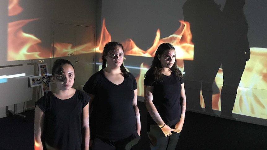 Three Indigenous high school students stand in front of a projection while filming a video.