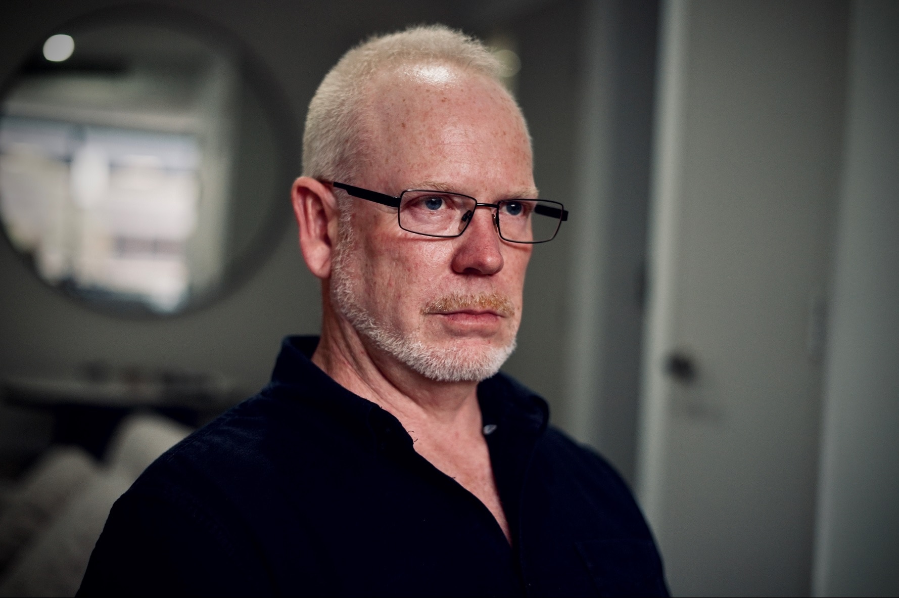 An older Caucasian male, wearing glasses and a black buttoned shirt.