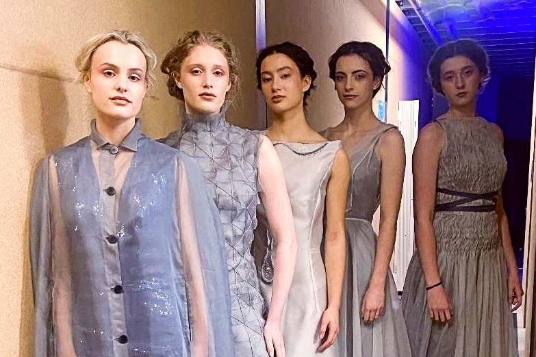 Five female models in light grey organza dresses stand backstage 