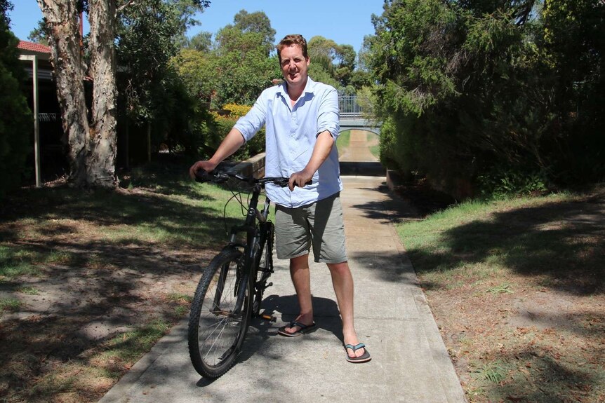 A man wearing shorts and a collared shorts stands in a laneway with his bike