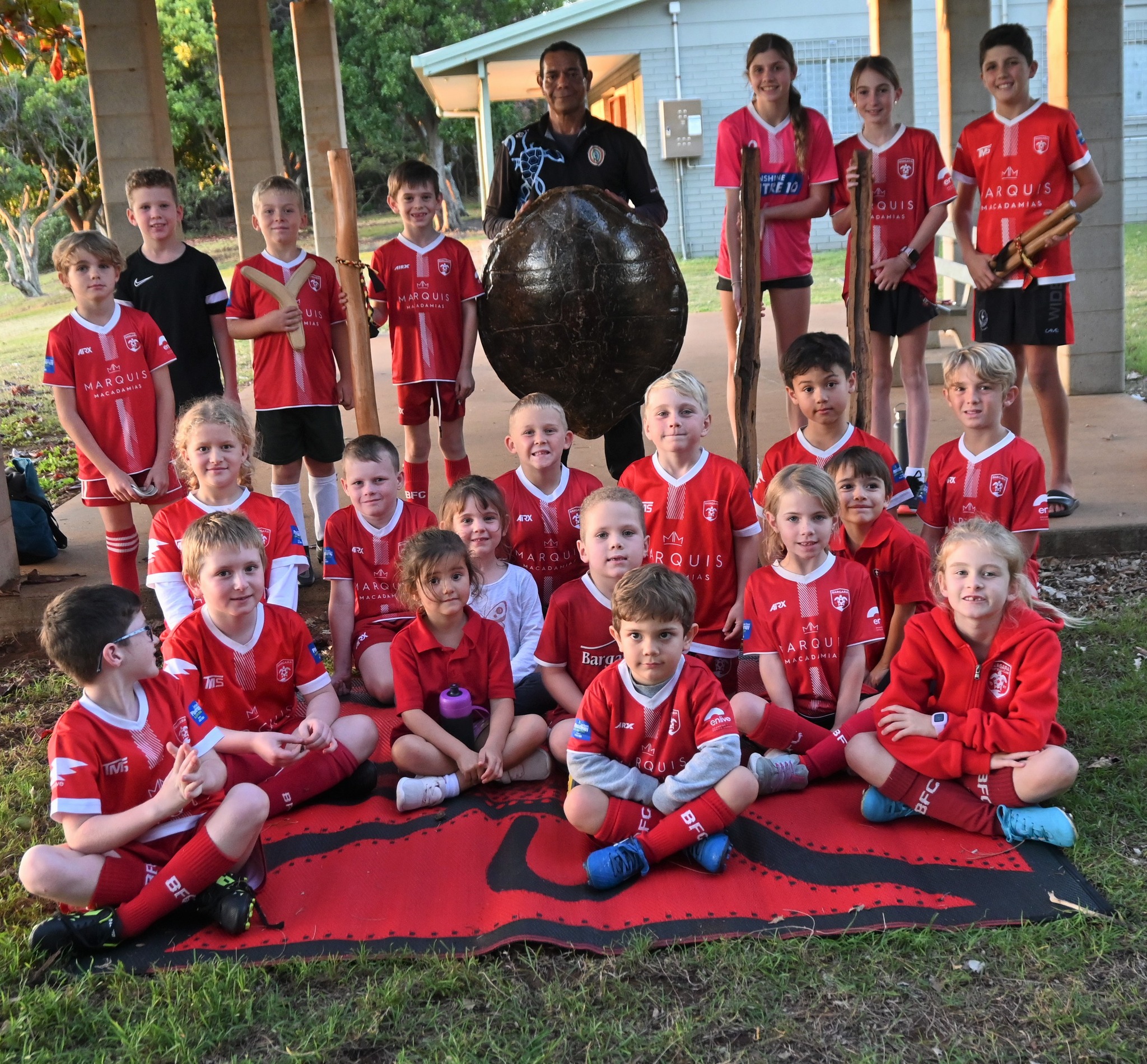 A large children's' soccer team sit with a First Australian educator standing behind them.