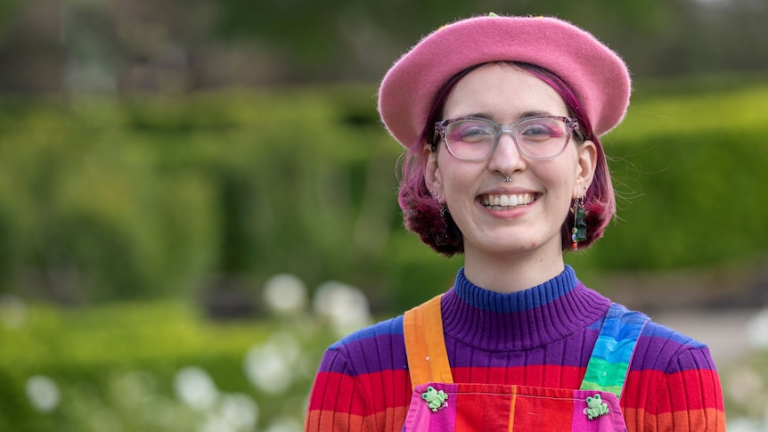 A young woman with pink beret, pink eyeshadow, pink hair, rainbow jumper smiles at the camera.