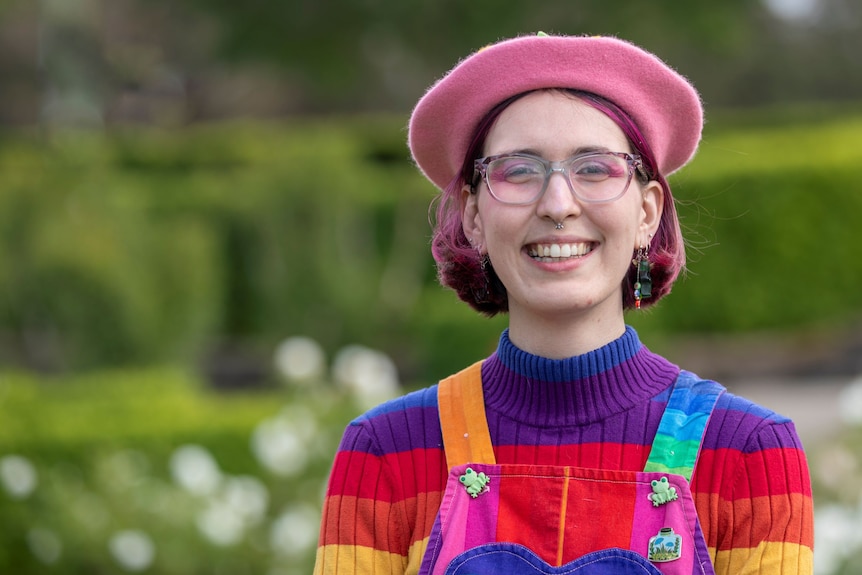 A young woman with pink beret, pink eyeshadow, pink hair, rainbow jumper smiles at the camera.