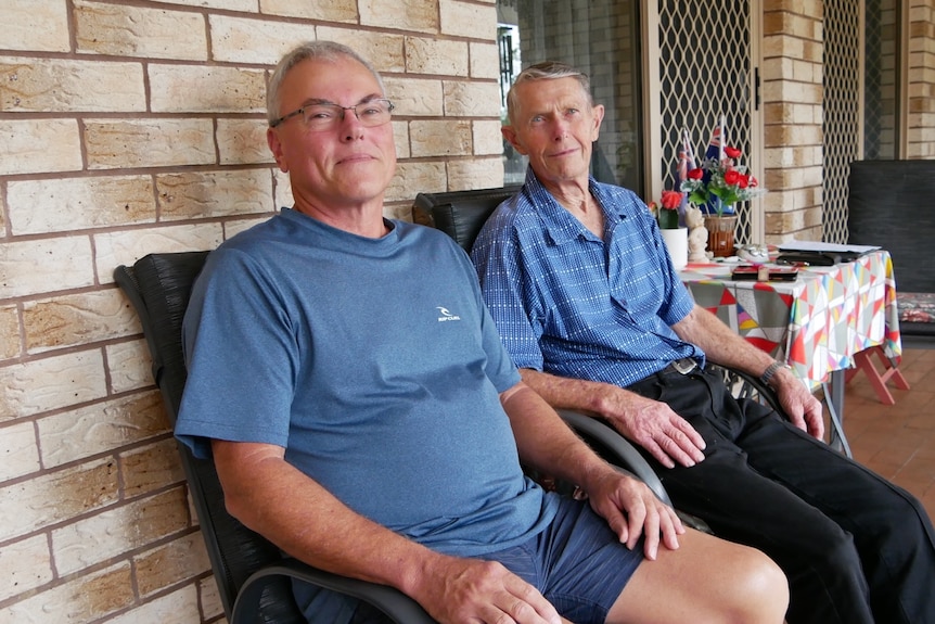 Two men sit on chairs outside a brick house.