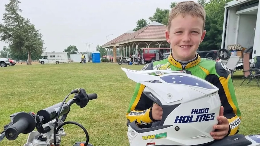 A young boy stands with his motorcycle helmet that has his name Hugo Holmes on it