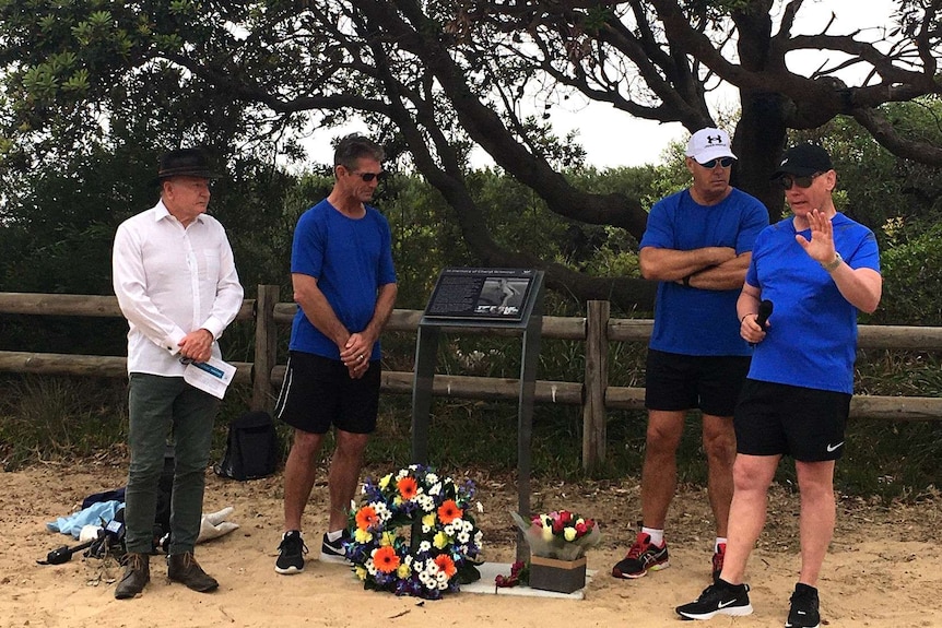 Four men, three dressed in matching shirts, standing around a plaque adorned with flowers in front of bushland at a beach.