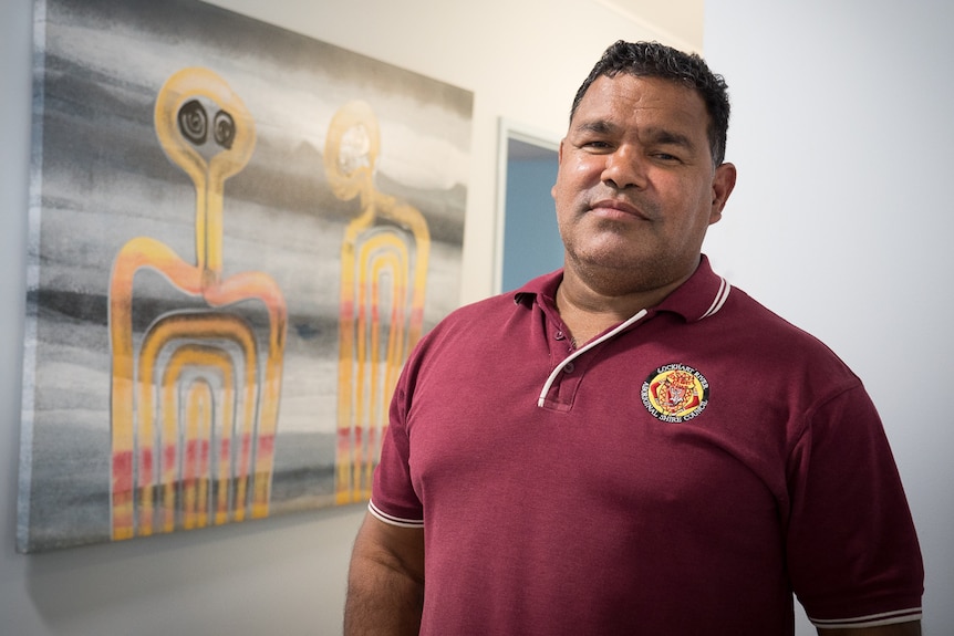 Councillor Wayne Butcher, Mayor of the Lockhart River Aboriginal Shire Council, stands in front of a Fiona Omeenyo artwork.