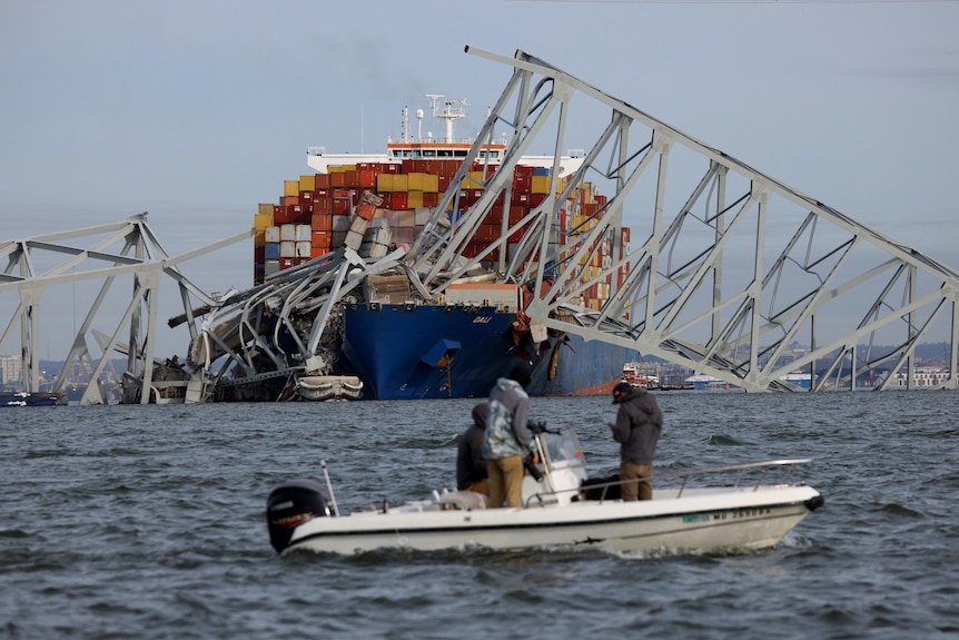 A small motorboat in front of a collapsed bridge with a large container ship underneath it