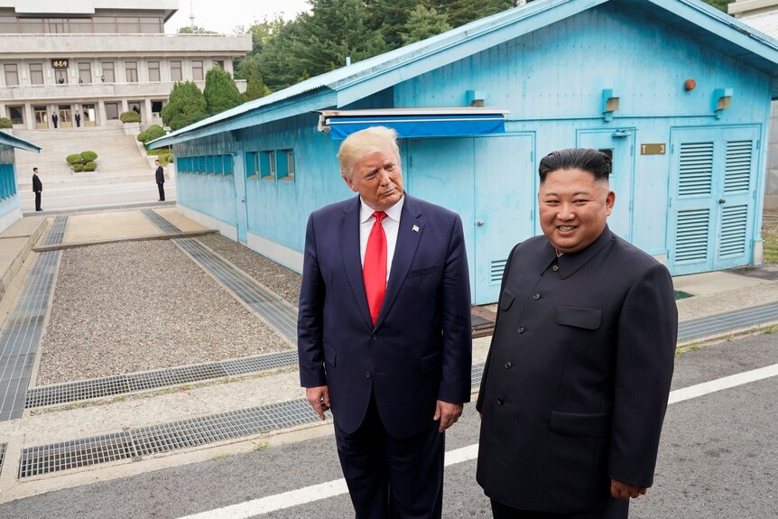 Donald Trump stands beside Kim Jong Un with a blue building in the background and walkway
