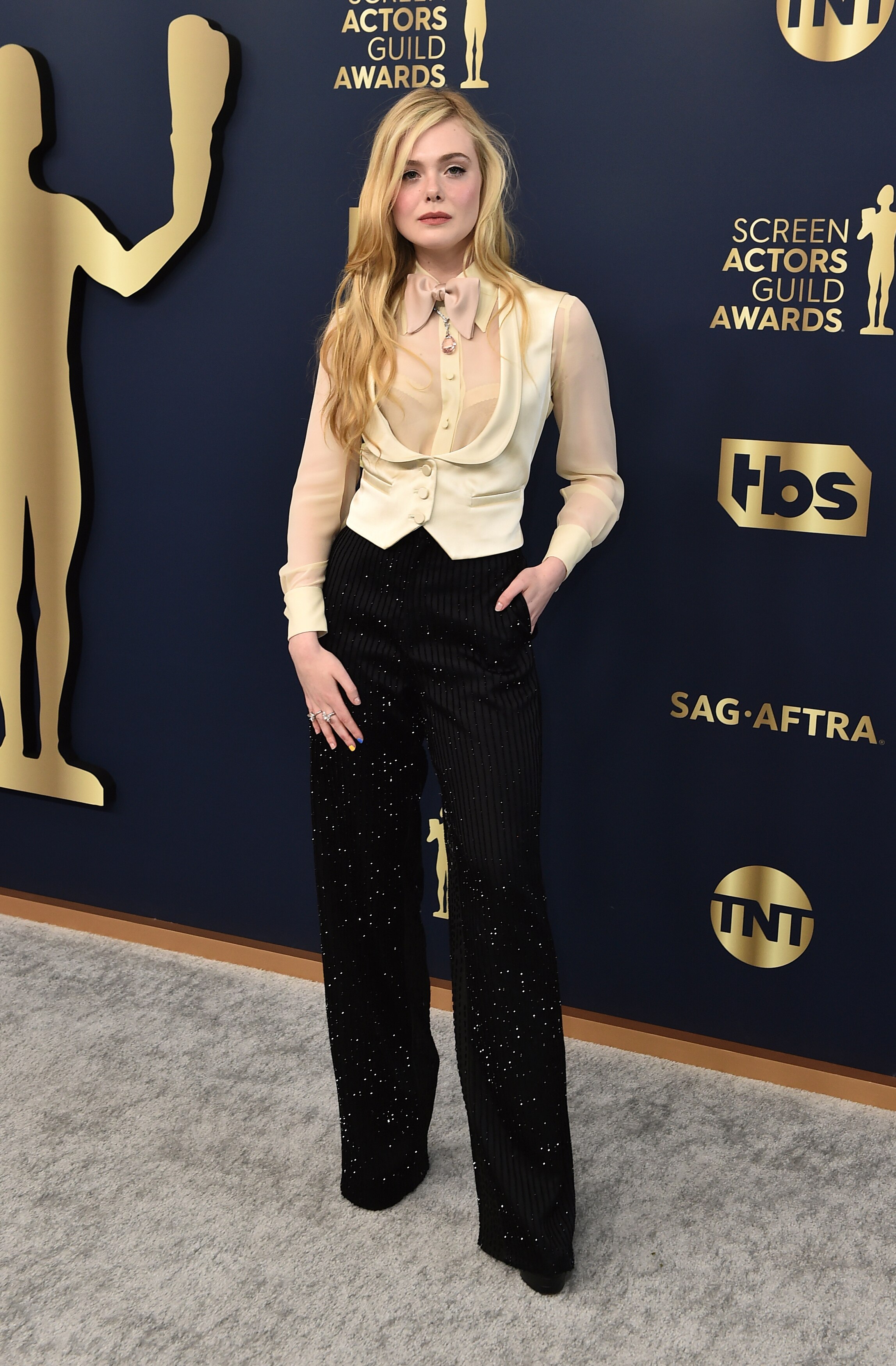 Elle Fanning wearing a white silk waistcoat over a blush button up shirt and sequinned balck trousers. 